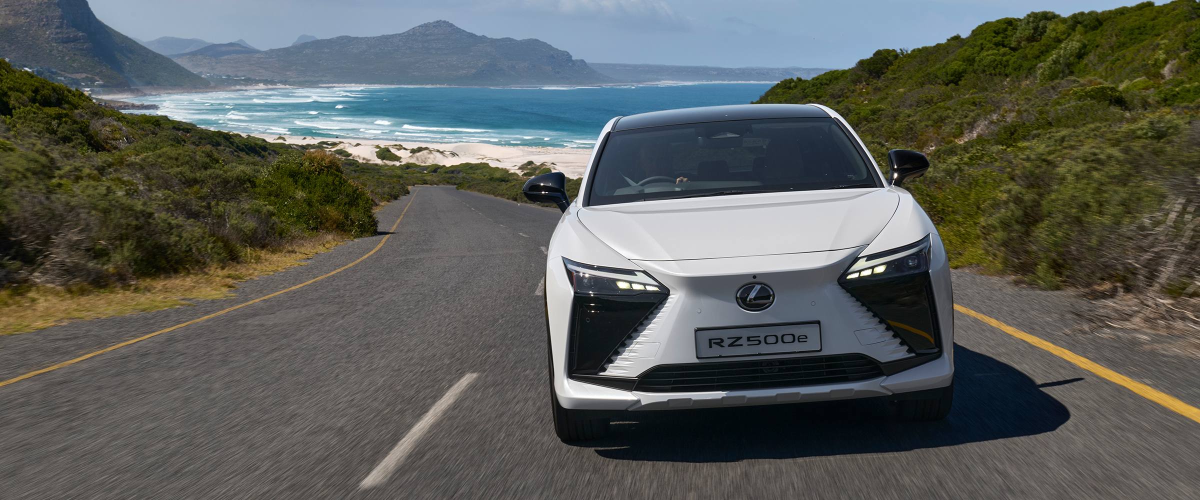 A white Lexus RZ 500e driving along a tree-lined road